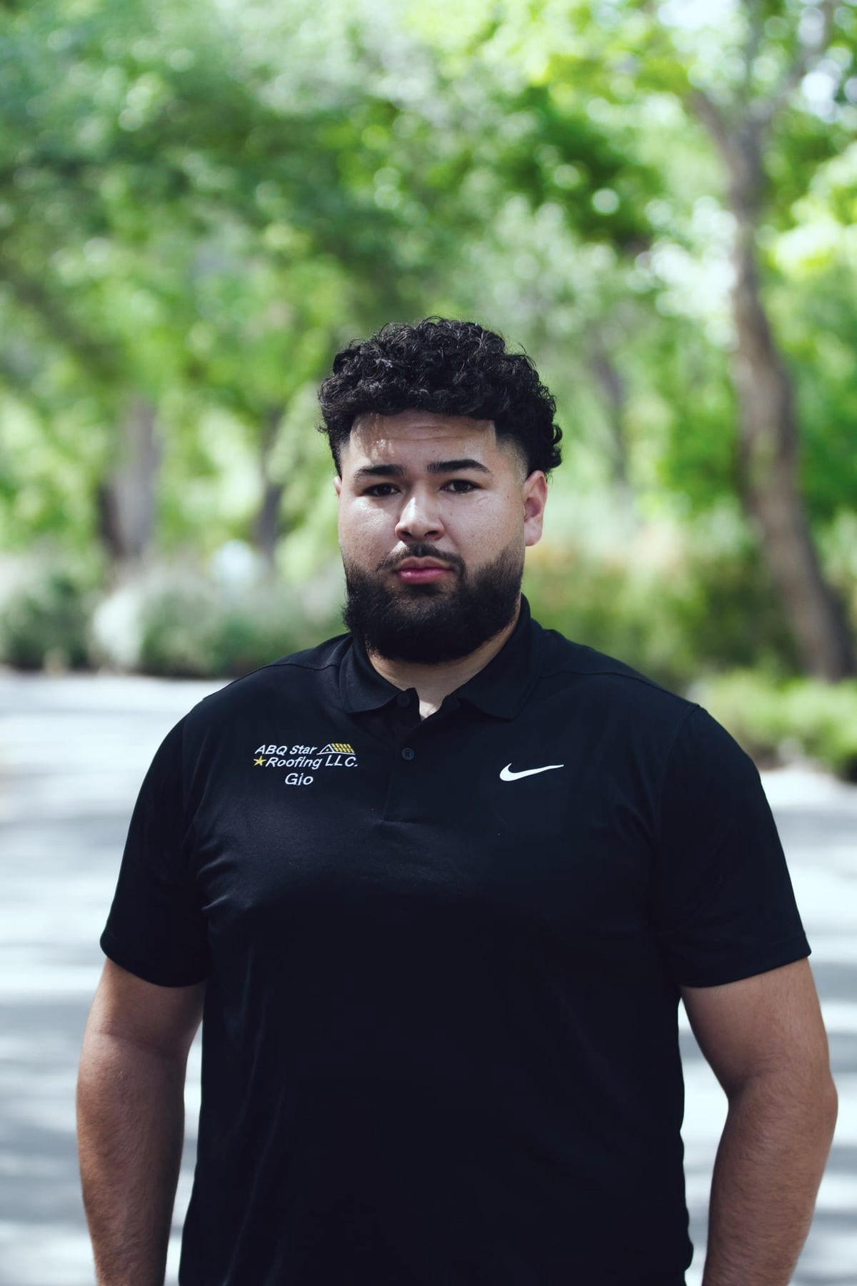 A member of the ABQ Star Roofing crew photographed outdoors against blurred green foliage. His black polo shirt is embroidered with the company's star-and-gable logo and the name Gio.