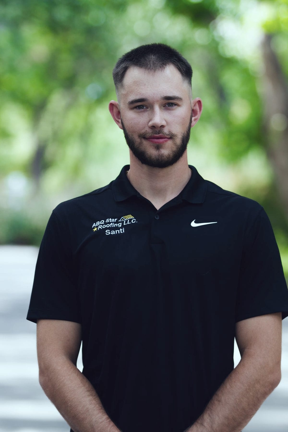 A member of the ABQ Star Roofing crew standing outdoors against blurred green foliage, hands clasped in front of him. His black polo shirt is embroidered with the company's star-and-gable logo and the name Santi.