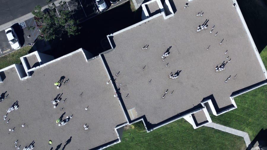 A large flat roof across two building wings, seen straight down from above. Dozens of plumbing vents and roof penetrations are spread over the surface, and workers in high-visibility vests are on the roof.