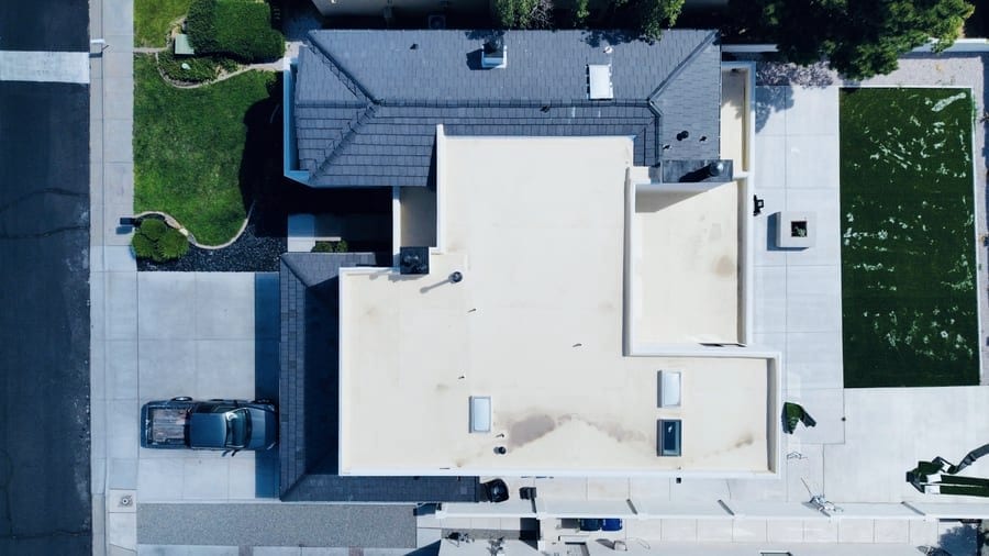 An overhead view of a home where a large cream-colored flat roof meets dark charcoal pitched shingle sections. Three skylights are set into the flat area, which is bounded by a low parapet.