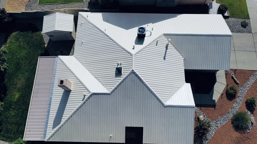 An overhead view of a white standing-seam metal roof on a single-story home. The panels run down four hip planes into the valleys, and an evaporative cooler and several plumbing vents sit on the slopes.