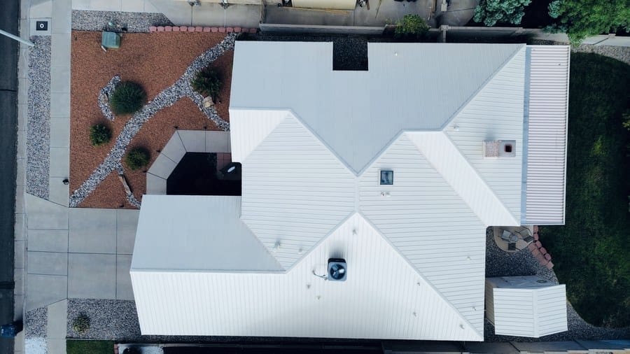 A white standing-seam metal roof seen straight down from above. The ribbed panels meet along the hips and valleys, and a chimney with a terracotta flue liner sits at the edge of one plane.