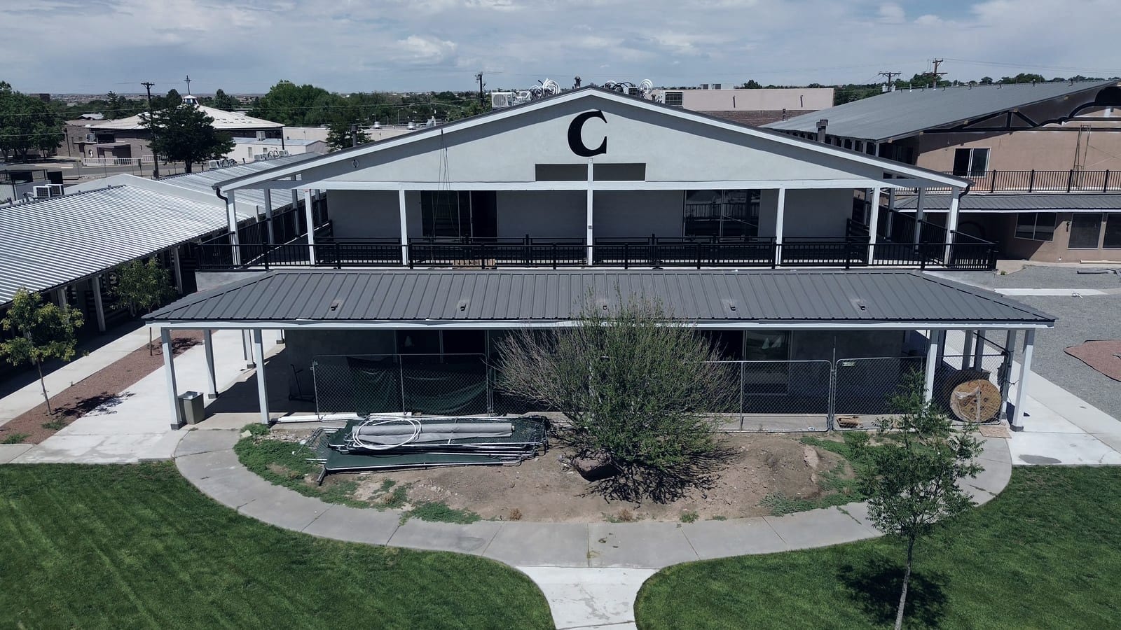 A two-story building with a dark gray standing-seam metal roof, photographed from the air. Matching metal roofs run along the covered walkways on either side, and a lawn and concrete paths curve around the front.