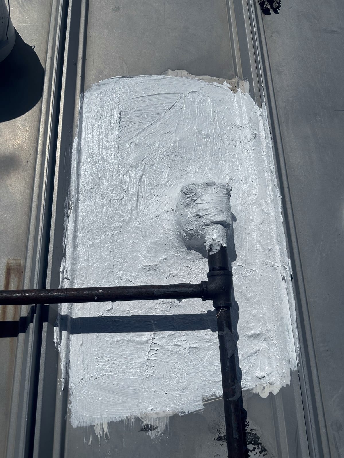 The same pipe penetration after resealing, covered in a smooth, unbroken white coating that laps up onto the pipe base. The rusted pipe and tee fitting above it are unchanged.