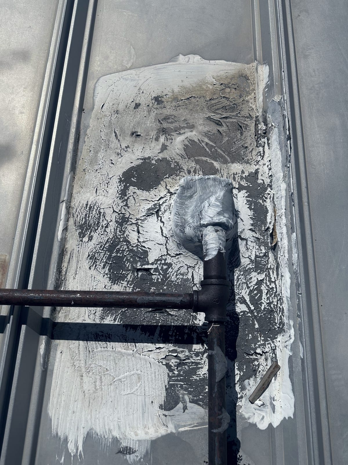 A pipe penetration on a metal roof where the old sealant has dried out and cracked into a network of splits, with bare gray substrate showing through in places. A rusted pipe and tee fitting cross the frame above it.