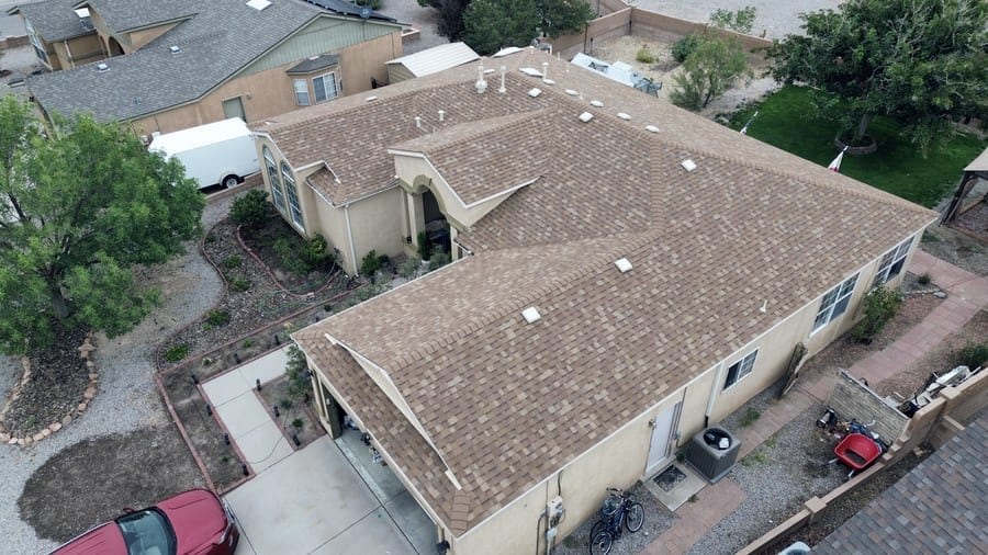 A single-story stucco home with a tan architectural shingle roof, seen from the air above the front of the house. Skylights and plumbing vents are set into the slopes, and an arched entry portico faces the driveway.