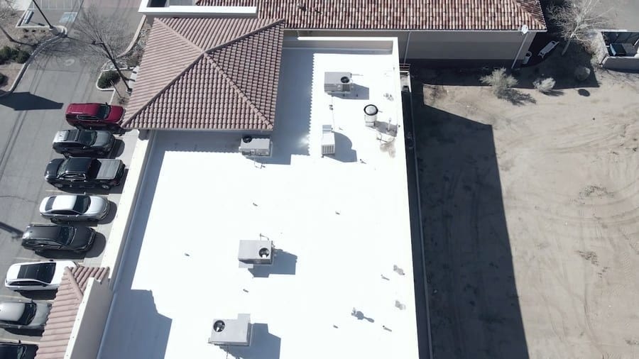 A white membrane flat roof meeting a terracotta barrel-tile pitched roof, photographed straight down. Rooftop mechanical units sit on the white section, and cars are parked along the building's edge.
