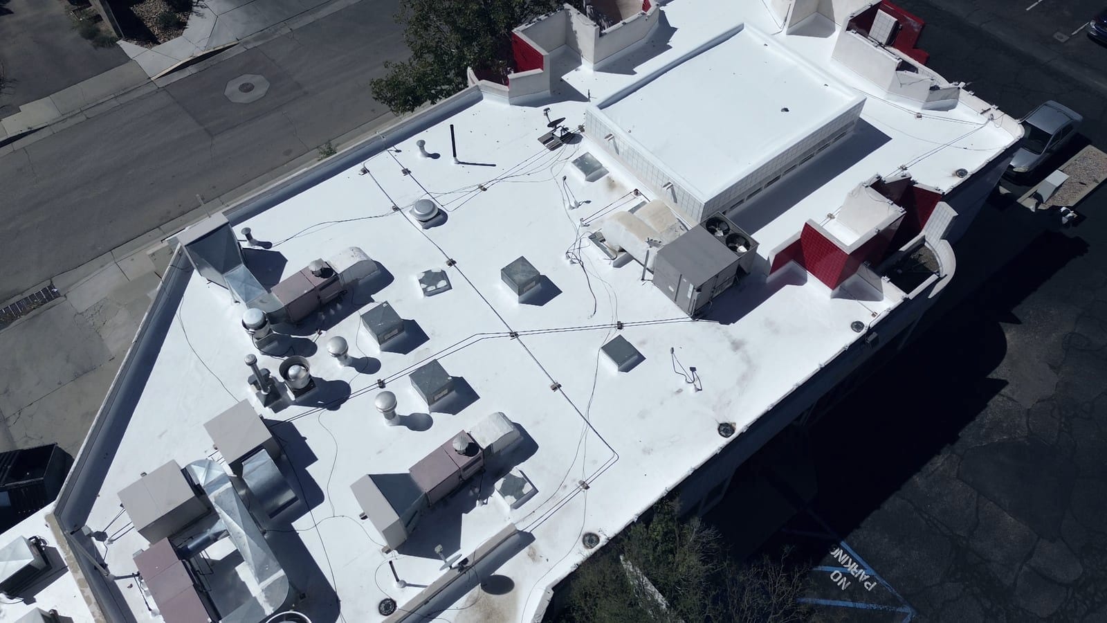 A bright white membrane roof on a commercial building, seen from above. Rooftop air-conditioning units, ducts and vents are spread across the surface, and red tiled parapet walls enclose the far edge.